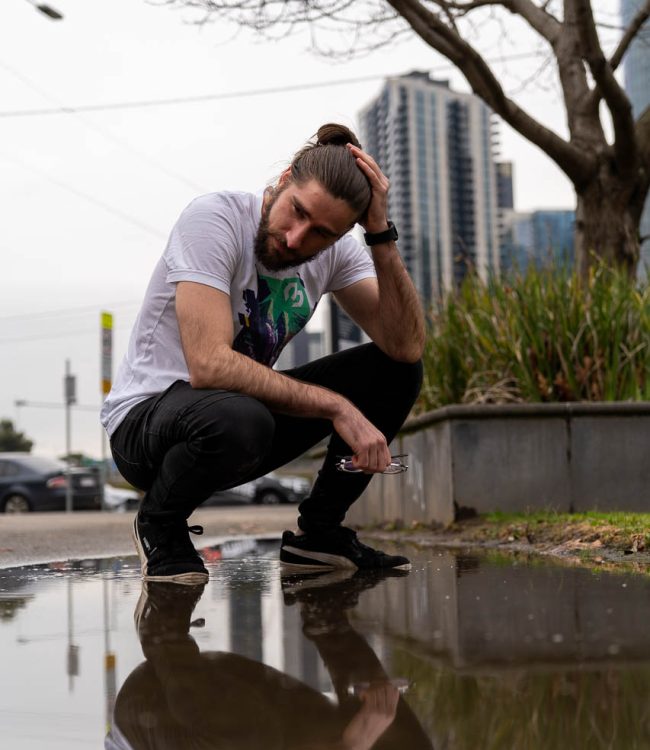 Mike Bello crouching over a puddle in the city wearing a white B-Side T-Shirt